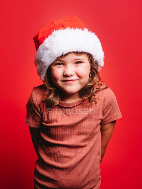 Adorable petite fille en vêtements décontractés et chapeau de Père Noël faire des visages tout en se tenant sur fond rouge et en regardant la caméra — Photo de stock