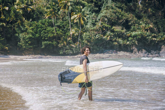 Jovem atleta do sexo masculino com cabelos cacheados e tatuagens em sportswear molhado segurando prancha de surf enquanto olha para a câmera na água — Fotografia de Stock