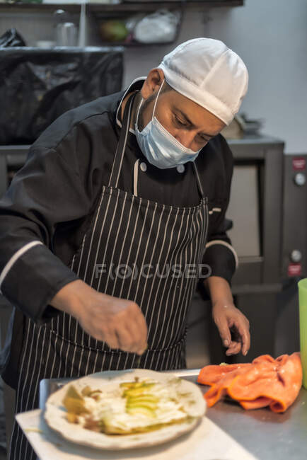 Middle aged ethnic male cook in uniform and protective mask salting fresh avocado slices on stuffed pasta in restaurant kitchen — Stock Photo