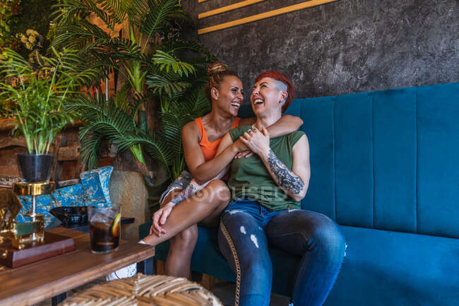 Zufriedene junge tätowierte Frau mit Mohawk und Getränk umarmt lesbische Freundin, während sie sich auf der Couch im Haus anschaut — Stockfoto