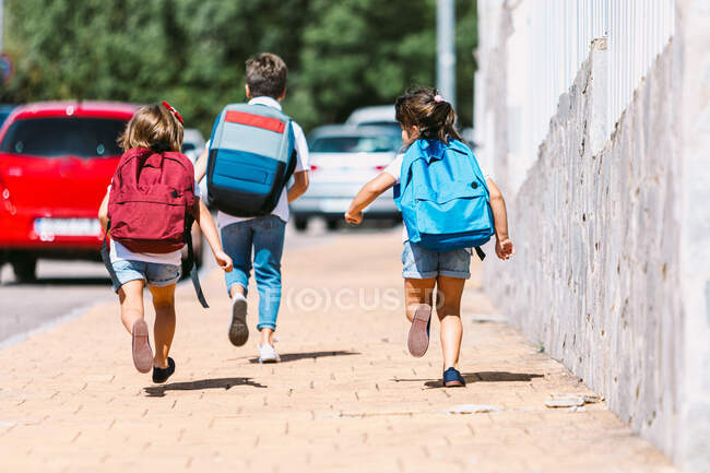 Vista posterior de los escolares anónimos con mochilas corriendo en la pasarela de azulejos en la ciudad soleada sobre un fondo borroso - foto de stock