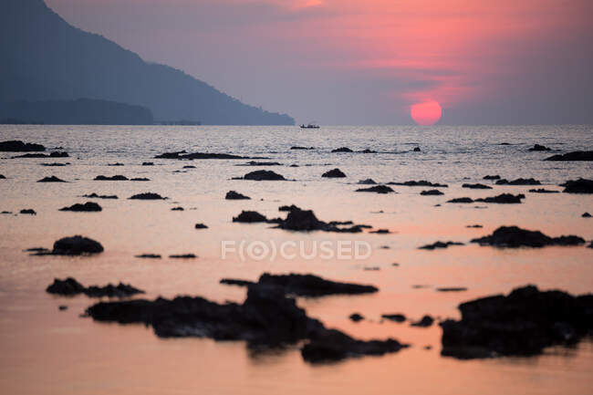 Wide coast with seaweeds of rippling sea against hill and red sun at sundown in Malaysia — Stock Photo