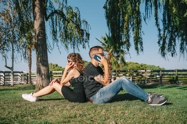 Вид збоку татуйованого чоловіка з дівчиною в сонцезахисних окулярах, що розмовляють на мобільних телефонах, сидячи назад на лузі в літньому парку — стокове фото