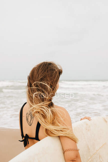 Vista trasera de una joven deportista irreconocible en traje de baño con tabla de surf mirando hacia la costa arenosa contra el océano tormentoso - foto de stock