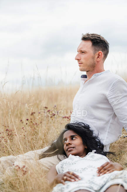 Dreamy Indian woman lying on thigh of male beloved while looking away on meadow under cloudy sky in fall — Stock Photo