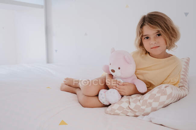 Criança gentil abraçando urso macio enquanto olha para a câmera e deitado na cama em casa — Fotografia de Stock
