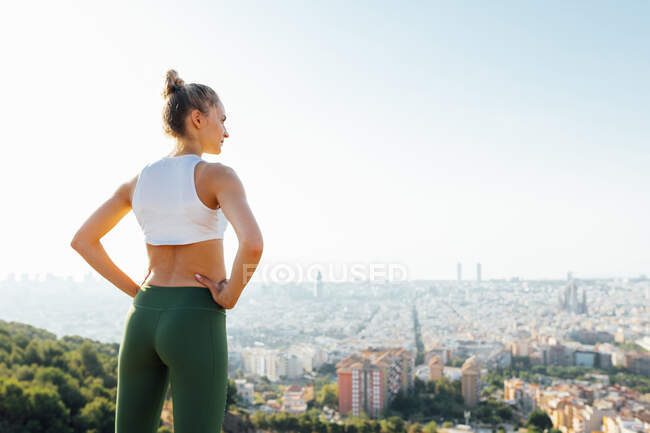 Vista posterior del atleta femenino en forma con las manos en la cintura admirando la ciudad de verano a la luz del sol - foto de stock
