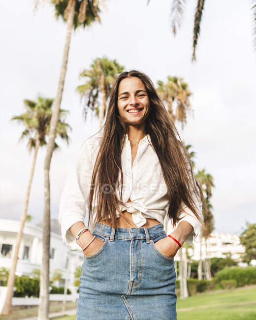Contenu adolescente aux cheveux longs en jupe en denim et chemise blanche regardant la caméra sur la prairie à Tenerife en Espagne — Photo de stock