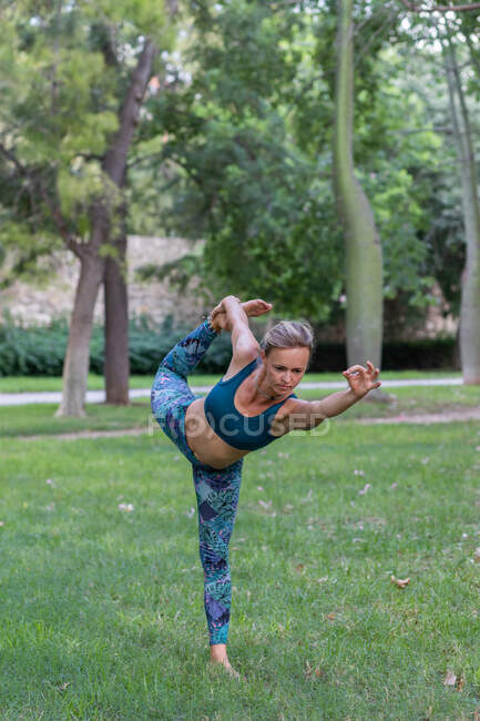 Full body of concentrated female in activewear doing Dandayamana Dhanurasana during yoga session in park in daytime — Stock Photo