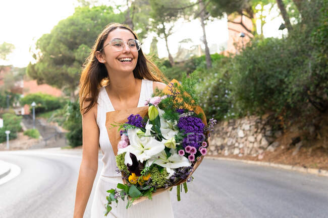 Amical jeune femme dans des lunettes avec bouquet floral en fleurs regardant loin sur la route de la ville — Photo de stock