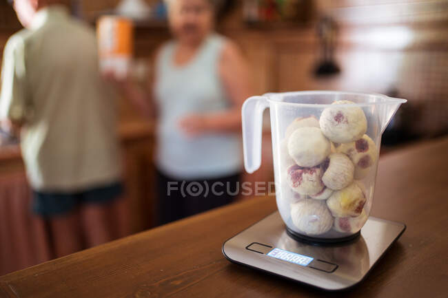 Ripe figs in pitcher on weighing scales with display on table against crop anonymous partners in kitchen — Stock Photo