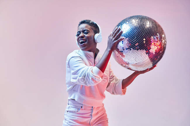Cool ethnic cheerful woman with short hair in wireless headset and trendy clothes dancing hip hop with open mouth and eyes closed holding glitter ball on violet background — Stock Photo