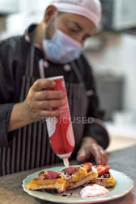 Crop etnico maschio cuoco in maschera sterile decorare deliziosi waffle viennesi con panna montata da bottiglia in cucina ristorante — Foto stock