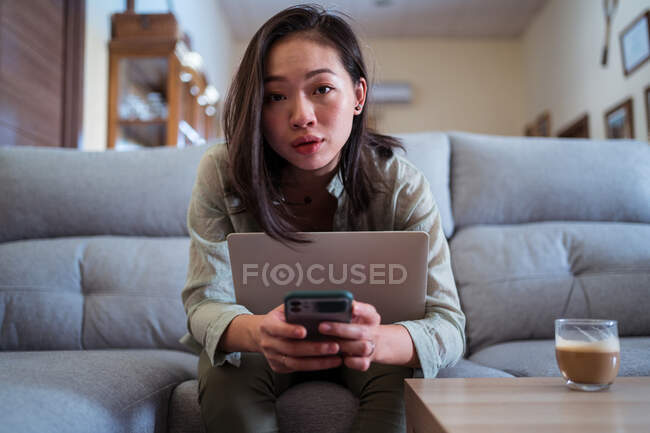 Joven mujer étnica con teléfono celular y netbook mirando a la cámara en el sofá contra el capuchino en la habitación de la casa - foto de stock
