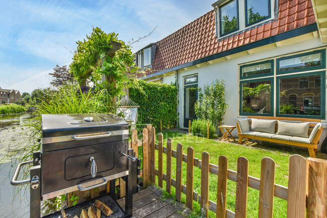 Barbecue fermé placé près d'une clôture en bois sur la terrasse avec de l'herbe verte et des plantes près du chalet moderne en été — Photo de stock