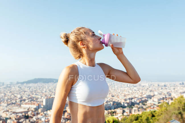 Тонкая спортсменка с рукой на бедре и закрытыми глазами пьет воду из бутылки во время перерыва от тренировки в городе — стоковое фото