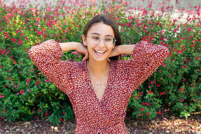 Joyeux jeune femme en robe rouge debout avec les bras levés et riant contre la floraison buisson dans la journée — Photo de stock
