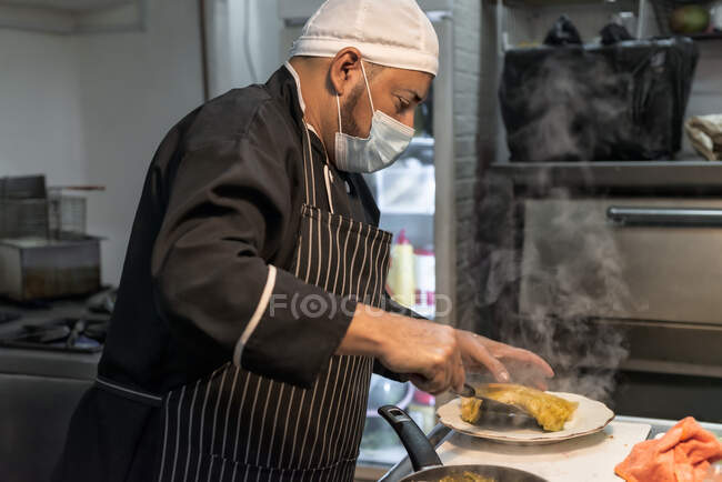 Vue latérale du cuisinier masculin ethnique mature en masque stérile avec spatule mettant des pâtes chaudes sur l'assiette dans la cuisine du restaurant — Photo de stock
