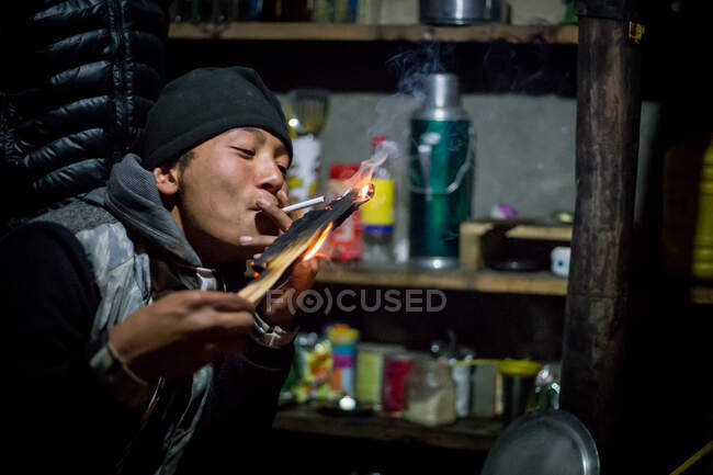 NEPAL, HIMALAYAS - 6 DE NOVIEMBRE DE 2015 Joven hombre étnico con ropa casual y sombrero fumando cigarrillo mientras está sentado en la mesa en la cafetería y mirando a la cámara - foto de stock
