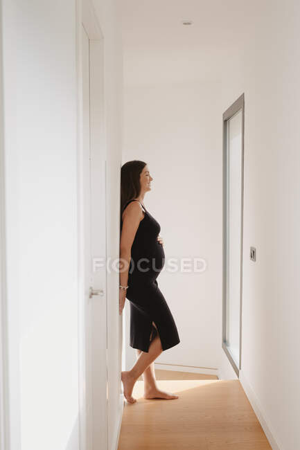 Mujer adulta soñadora expectante con los ojos cerrados acariciando la barriga mientras está de pie en la casa en el día soleado - foto de stock