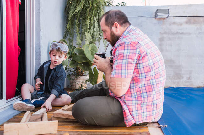 Hipster papá bebiendo té de hierbas de calabaza calabaza contra hijo trabajando con madera - foto de stock