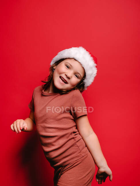 Очаровательная счастливая маленькая девочка в повседневной одежде и в шляпе Санты, улыбающаяся стоя на красном фоне и глядя в камеру — стоковое фото