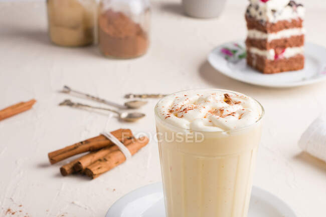 Glas Milchpunsch mit Zimtpulver auf geschlagenem Eiweiß gegen Kuchenstück auf Cafeteria-Tisch auf hellem Hintergrund — Stockfoto
