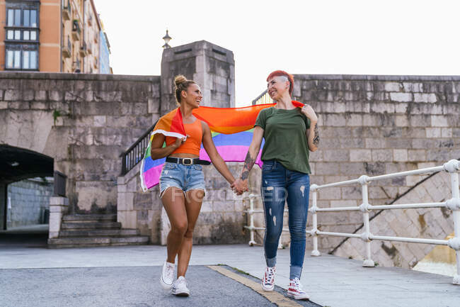 Joyeux jeunes copines homosexuelles fraîches avec drapeau LGBTQ tenant la main tout en se regardant et se promenant sur le trottoir urbain — Photo de stock