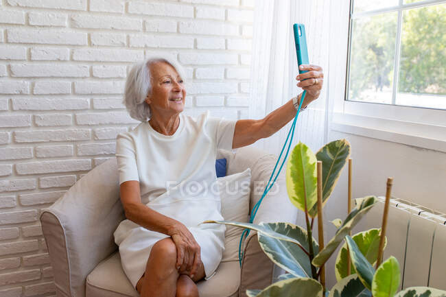 Smiling senior female sitting in armchair and speaking on video call via mobile phone at home — Stock Photo