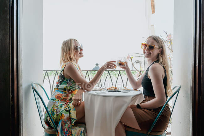 Vista lateral de amigas felices almorzando en la mesa y tintineando vasos de cócteles en el balcón - foto de stock