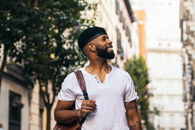 Smiling trendy young African American hipster male in beret and rings looking away in town on blurred background — Stock Photo
