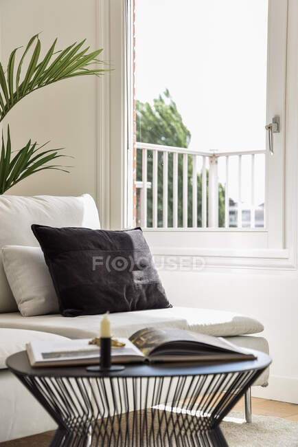 Sofa with soft cushions placed near table with opened magazine and candle near window in living room — Stock Photo