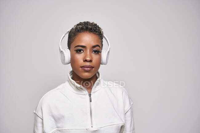 Mujer étnica seria en ropa de moda escuchando música en auriculares inalámbricos mirando a la cámara - foto de stock