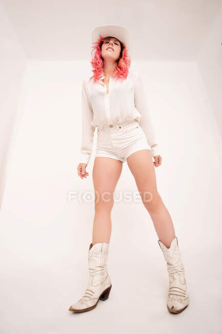 Von unten steht eine junge Frau mit pinkfarbenen Haaren in stylischer weißer Kleidung und Hut im hellen Raum — Stockfoto