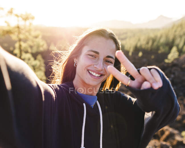Candid female teen in sweatshirt taking selfie with smartphone while demonstrating victory gesture looking at camera against mount in Tenerife in backlit — Stock Photo