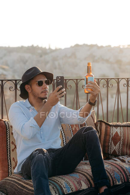 Moderno hombre con ropa elegante y gafas de sol sentado en cojines y tomando una foto de botella de vidrio de cerveza en el teléfono celular en el bar terraza en Capadocia, Turquía - foto de stock