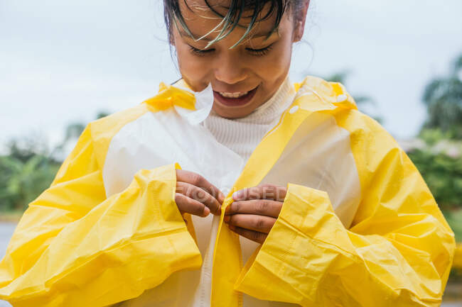 Crop content ethnic child buttoning up white and yellow slicker in rainy weather on blurred background — Stock Photo