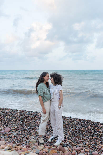 Junge lesbische Freundinnen in Freizeitkleidung umarmen sich, während sie sich an der Meeresküste unter wolkenverhangenem Himmel anschauen — Stockfoto