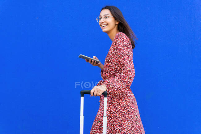 Vista lateral de la mujer positiva en vestido largo rojo caminando con el equipaje mientras navega en el teléfono inteligente en la calle contra la pared azul en el día - foto de stock