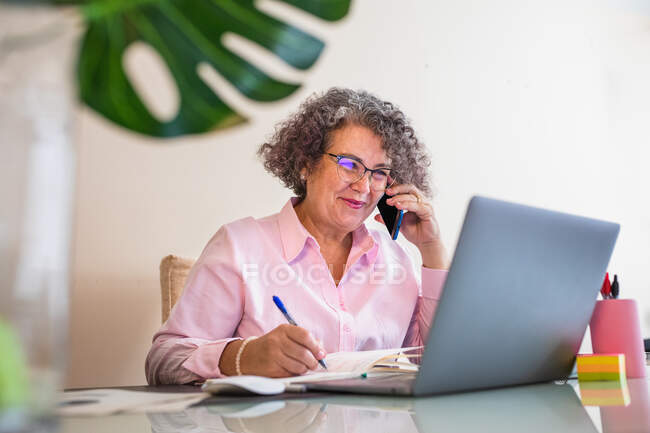Cheerful elderly female entrepreneur in eyewear with pen speaking on cellphone against netbook on table in workspace — Stock Photo