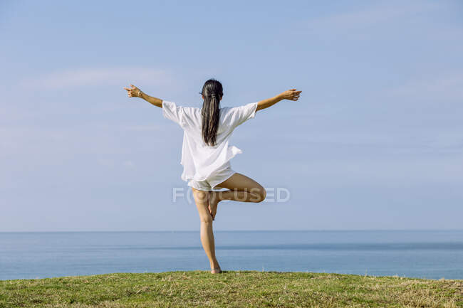 Back view of anonymous barefoot female performing Vrikshasana pose with outstretched arms during yoga practice on grass shore against ocean — Stock Photo