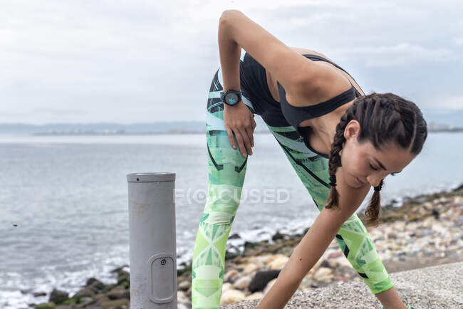 Focused female athlete doing side lunge exercise and stretching legs while warming up during training on embankment near sea — Stock Photo