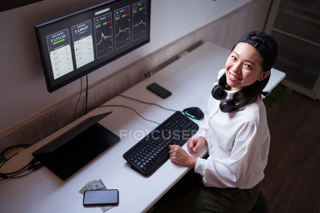 Alto ángulo alegre mujer asiática que trabaja en el ordenador con gráficos que muestran la dinámica de los cambios en el valor de la criptomoneda en el lugar de trabajo conveniente - foto de stock