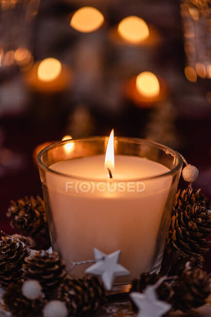 Пламенная свеча в стеклянном держателе украшена конусами, прижатыми к блестящим огням для празднования Рождества — стоковое фото