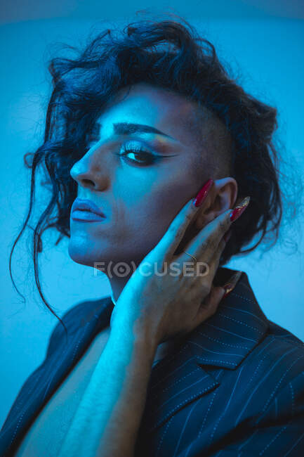 Jeune modèle masculin transsexuel avec maquillage dans une veste élégante regardant la caméra sur fond bleu — Photo de stock