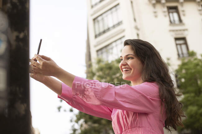 From below cheerful female standing on sidewalk and taking picture on mobile phone in city — Stock Photo
