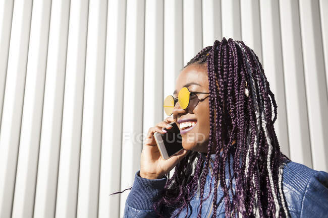 Cheerful stylish young African American female with Afro braids wearing trendy jacket and sunglasses speaking on mobile phone while standing near wall of urban building — Stock Photo