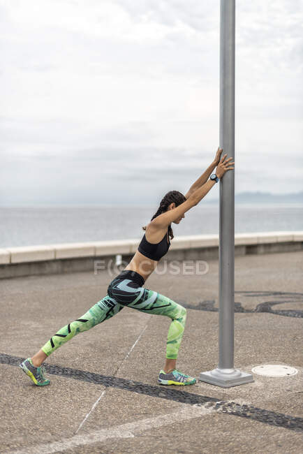 Vista lateral do atlético feminino corredor apoiando-se no pilar e esticar as pernas enquanto se aquece no aterro durante o treino no verão — Fotografia de Stock