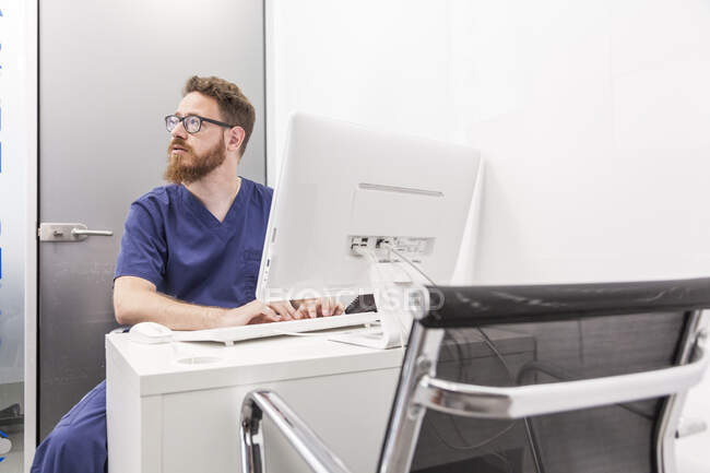 Homme en uniforme médical tapant sur le clavier de l'ordinateur tout en travaillant dans une clinique de soins de santé — Photo de stock