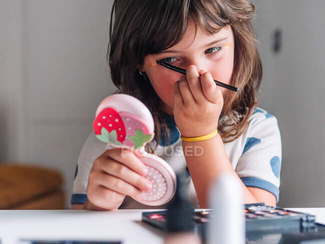 Criança com aplicador e espelho que compõem o rosto à mesa com produtos cosméticos variados em casa — Fotografia de Stock